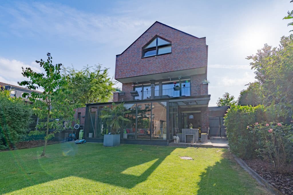 Ein zeitgemäß gestaltetes Haus mit großen Fenstern und überdachter Terrasse lädt zum Wohlfühlen ein. Umgeben von grüner Wiese, Bäumen und Sträuchern vereint es natürliche Materialien, modernes Design und behagliche Atmosphäre bei Sonnenlicht.