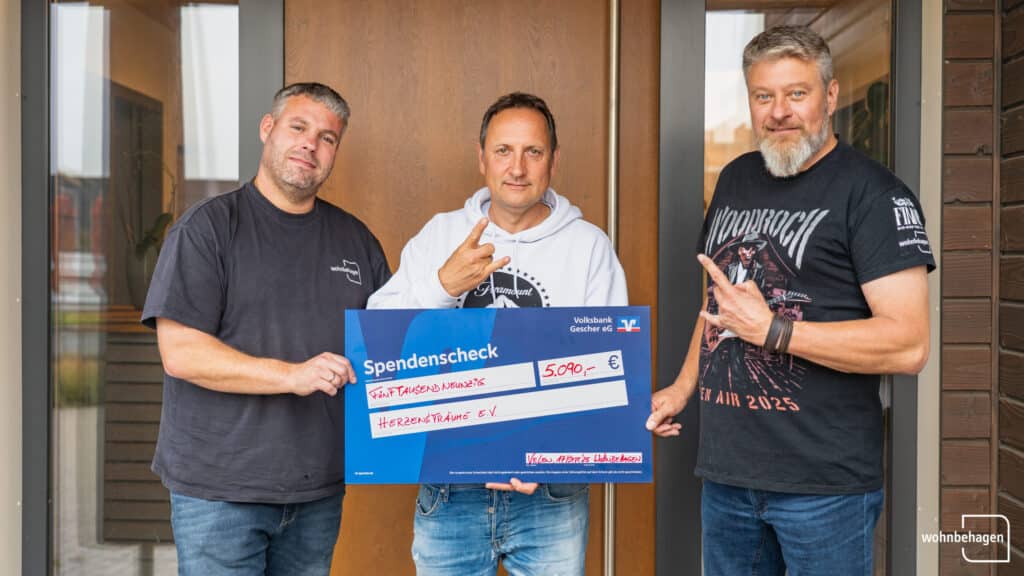 Drei Männer stehen gemeinsam vor einem freundlich gestalteten Hintergrund und halten einen großen Spendenscheck über 5090 Euro für Herzensträume e.V. bei einer WoodRock Spendenübergabe. Lächelnd zeigen zwei auf den Scheck, während einer gestisch begleitet.