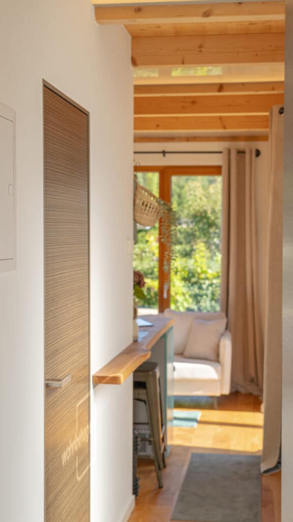 Ein heller Flur mit sichtbaren Holzbalken führt in einen behaglichen Wohnbereich. Ein weißer Armlehnsessel steht einladend am großen Fenster, das viel Tageslicht hereinlässt und den Blick ins Grüne öffnet - für ein Zuhause voller Wärme und Komfort.