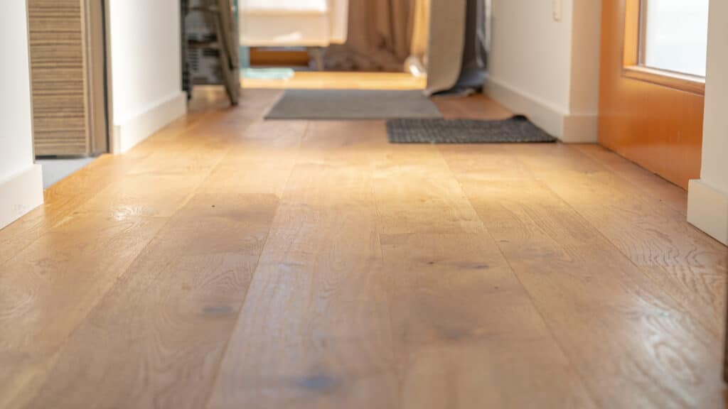 Ein heller Holzfußboden bringt Wärme in den Flur, während sanftes Licht durch die Fenster am Ende fällt und weiche Schatten auf dem Boden spielt. Eine kleine Ablage links und Türvorleger sorgen für einen einladenden Wohlfühlcharakter.