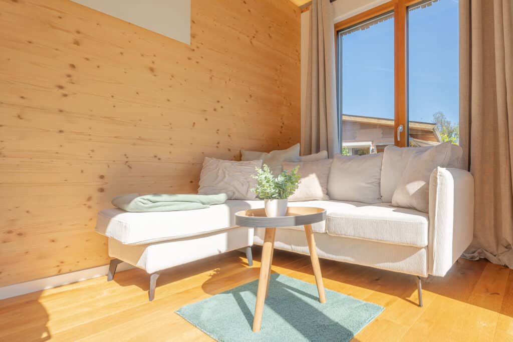 Heller Wohnraum mit einer Wand aus hellem Holz, einem gemütlichen weißen Ecksofa samt Kissen und grüner Decke sowie einem runden Holzcouchtisch mit Zimmerpflanze. Große Fenster mit beigen Vorhängen lassen sanftes Sonnenlicht herein.