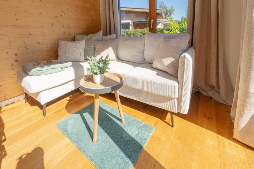 Eine helle Wohnzimmerecke mit einem weißen L-förmigen Sofa, neutralen Kissen und einer kuscheligen grünen Decke lädt zum Entspannen ein. Ein runder Holztisch mit einer Topfpflanze und Sonnenlicht auf dem Holzfußboden sorgen für natürliche Behaglichkeit.