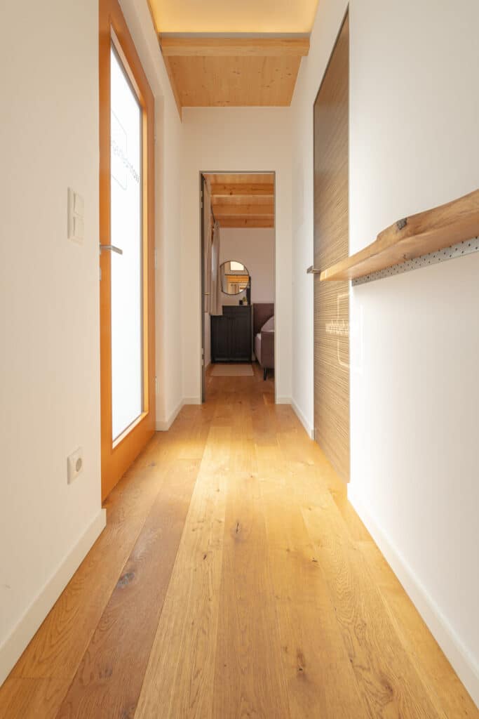 Ein heller Flur mit natürlichen Holzböden und weißen Wänden führt behaglich in ein Schlafzimmer. Links befindet sich eine Tür aus satiniertem Glas, rechts eine schlichte Holzablage. Sonnenlicht durchflutet den Raum und sorgt für wohnliche Wärme.