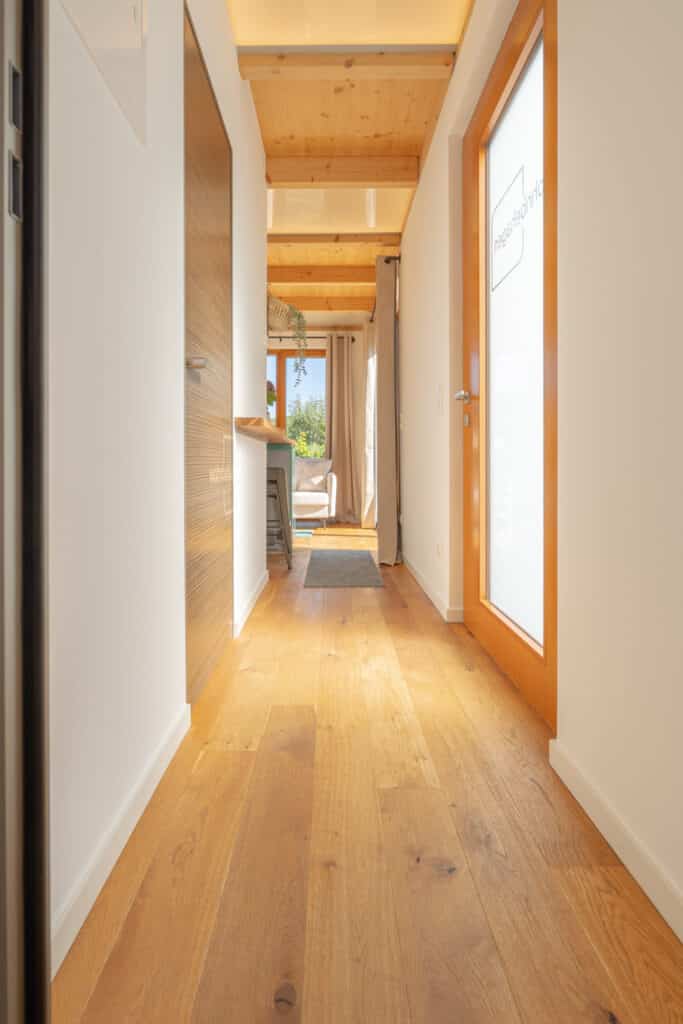 Eine einladende Flur mit hellen Holzböden und passender Holzdecke schafft eine warme Atmosphäre. Rechterhand verbindet eine Glastür mit satiniertem Einsatz moderne Eleganz und Privatsphäre, während links eine Holztür Natürlichkeit betont. Am Ende des Flurs fällt Sonnenlicht durch ein Fenster und sorgt für behagliches Wohngefühl.