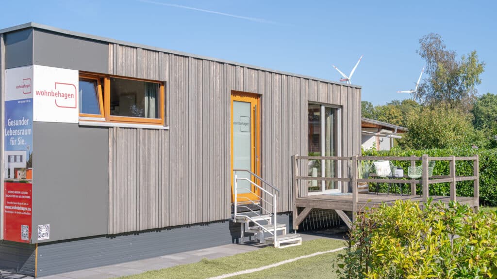 Ein modernes Tiny House aus Holz mit vertikaler Fassadenverkleidung, kleinem Fenster und Glastür öffnet sich zu einer erhöhten Terrasse mit Metallgeländer. Umgeben von viel Grün und unter blauem Himmel strahlt das Haus Wärme und Geborgenheit aus.