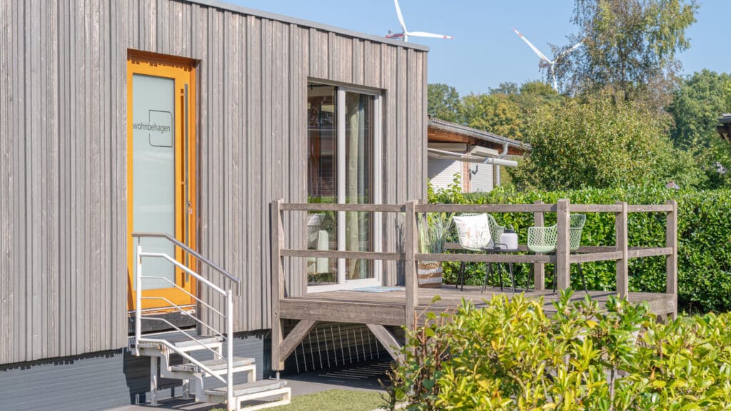 Ein modernes Tiny House aus Holz mit großen Fenstern lädt auf eine kleine Veranda mit gemütlichen Sitzgelegenheiten zum Verweilen ein. Metallstufen führen zur Haustür, umgeben von grüner Natur; im Hintergrund stehen Windräder für nachhaltige Energie.