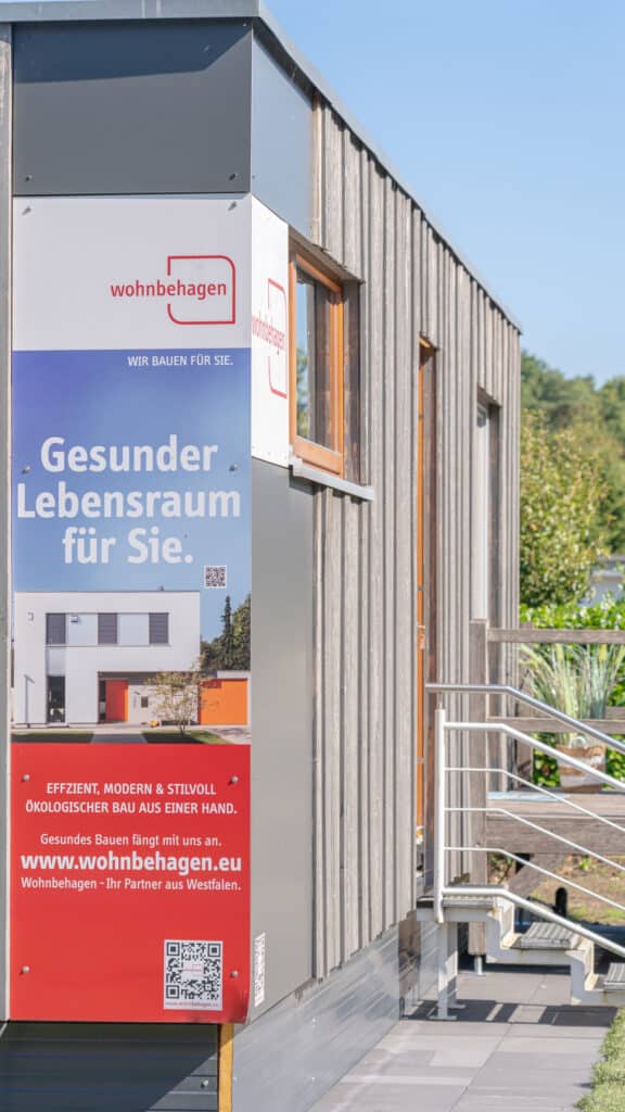 Ein modernes Holzhaus mit einem großen Schild "Gesunder Lebensraum für Sie" von wohnbehagen, das für nachhaltige Fertighäuser wirbt. Das Schild zeigt einen QR-Code, die Website und ein Bild eines weißen Hauses - Wohlfühlen und Qualität sichtbar.