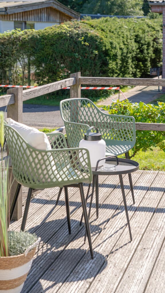 Zwei grüne Mesh-Patio-Stühle mit gemütlichen Kissen stehen einladend um einen kleinen, runden Tisch mit stilvoller weißer Laterne. Die Holzterrasse wird von grüner Bepflanzung und einem Zaun umrahmt - entspannter Rückzugsort im eigenen Zuhause.