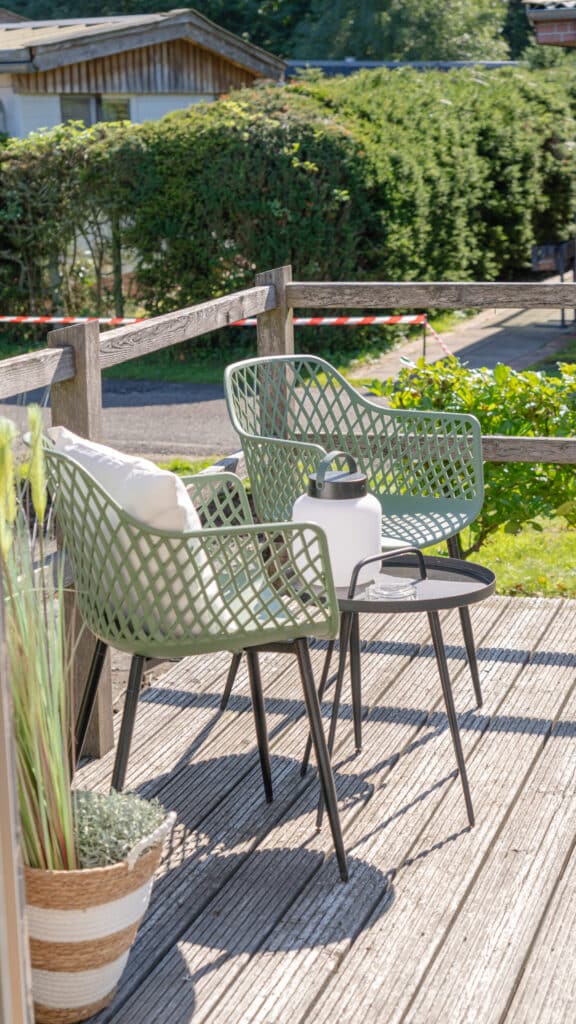 Auf einer Holzterrasse laden zwei grüne Gartenstühle mit weißen Kissen zum Entspannen ein. Ein kleiner, schwarzer Tisch mit stilvoller Laterne ergänzt die gemütliche Sitzgruppe und eröffnet den Blick auf eine grüne Hecke und einen sonnigen Garten.