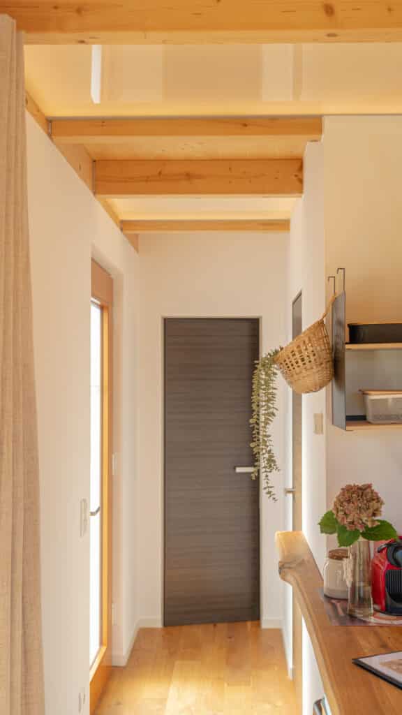 Ein heller, moderner Flur mit Holzfußboden und sichtbaren Deckenbalken, umgeben von weißen Wänden. Am Ende befindet sich eine dunkle Holztür. Regale mit Körben sowie eine Arbeitsfläche mit Pflanze und Küchenutensilien schaffen Behaglichkeit.