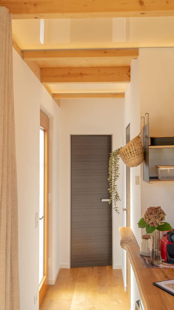 Ein einladender Flur mit warmen Holzböden und sichtbaren Deckenbalken führt zu einer geschlossenen, dunklen Tür. An der Wand hängt ein geflochtener Korb mit einer Pflanze, während Sonnenlicht durch Fenster und Glastür Behaglichkeit schafft.