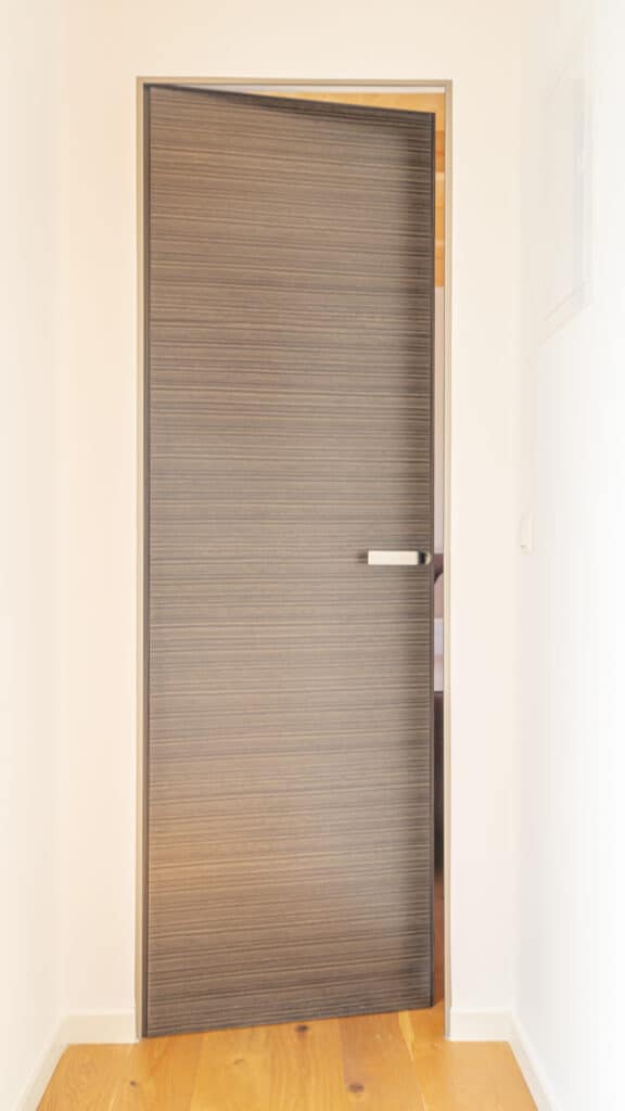 Eine halb geöffnete, moderne Holztür mit horizontaler Maserung begrüßt im hellen Flur und spiegelt zeitgemäßes Design sowie nachhaltige Bauweise wider. Der elegante, silberne Griff unterstreicht die warme Atmosphäre und hochwertige Ausführung.
