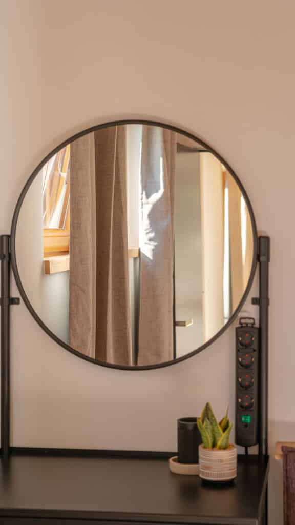 Ein runder Spiegel fängt das sanfte Licht eines sonnendurchfluteten Fensters und beige Vorhänge ein. Darunter sorgt ein schwarzer Schreibtisch mit Zimmerpflanze, kleiner Dose und schlichter Steckleiste für wohnliche Harmonie.