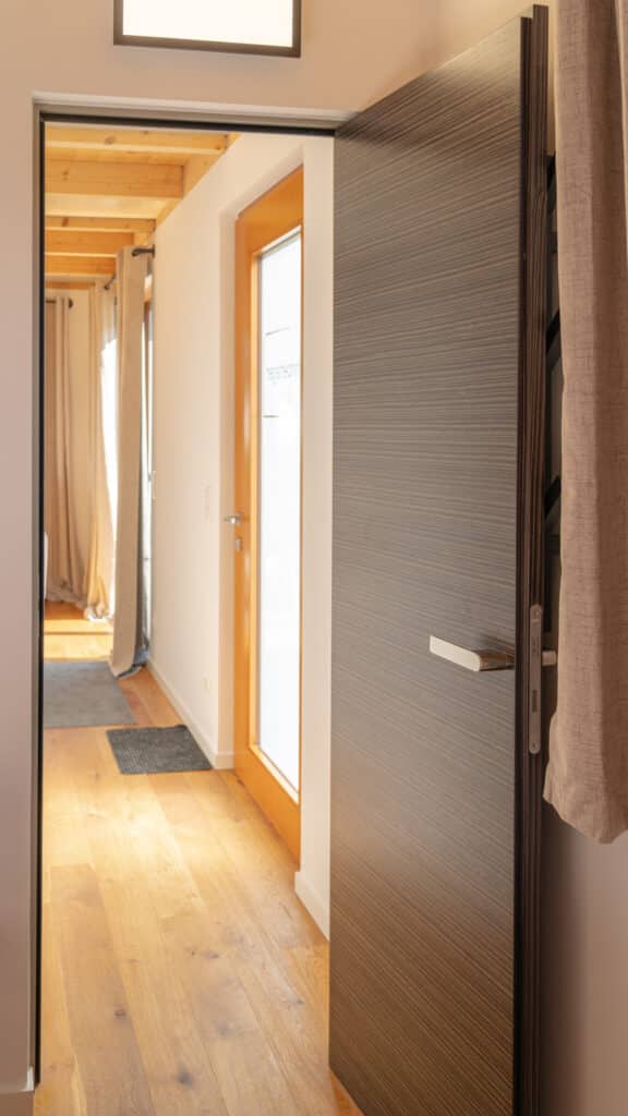 Ein moderner Flur mit warmen Holzböden und einer teils geöffneten dunklen Holztür lädt zum Wohlfühlen ein. Sonnenlicht fällt durch die verglaste Tür am Ende des Flurs und taucht die hellen Wände in ein behagliches Licht.