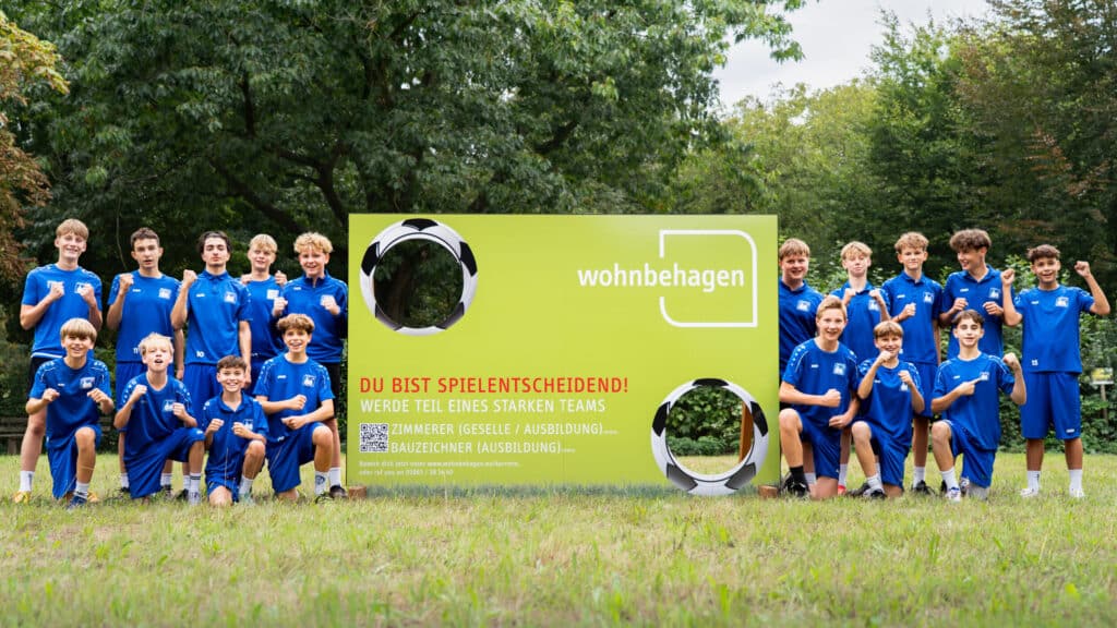 Eine Jugendfußballmannschaft in blauen Trikots steht draußen hinter einem großen grün-gelben Schild mit "wohnbehagen" und deutscher Schrift. Umgeben von Bäumen und Gras jubeln die Spieler freudig - voller Energie und Zusammenhalt.