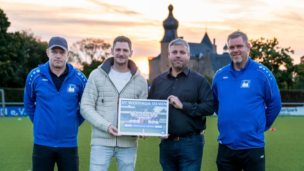 Vier Männer stehen entspannt auf einem Fußballfeld bei Sonnenuntergang zur Trikotübergabe von Westfalia Gemen. Zwei tragen blaue Trainingsanzüge, einer ist in Schwarz, einer hält im hellen Jackett ein eingerahmtes Teamfoto. Dahinter das charmante, historische Gebäude. Firma wohnbehagen hat die Trikots für die B2 Mannschaft von Westfalia Gemen gesponsort.