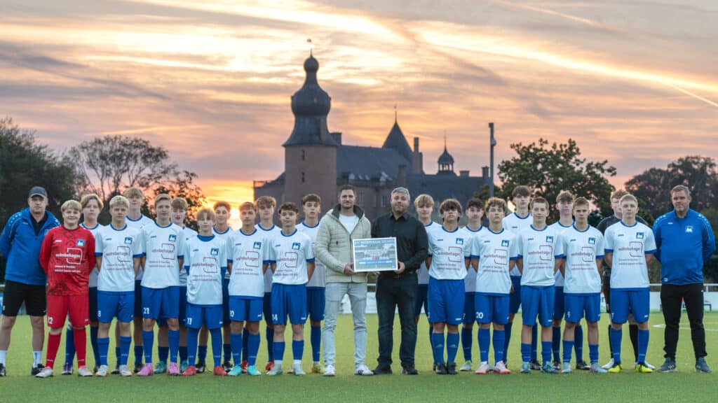 Zum Sonnenuntergang steht ein Jugendfußballteam in weißen und blauen Trikots gemeinsam für die Trikotübergabe Westfalia Gemen auf dem Platz. Umgeben von Trainern und Erwachsenen mit einem großen Scheck entsteht vor der Kulisse eines Schlosses eine Atmosphäre von Zusammenhalt, Freude und Geborgenheit - wie sie auch ein behagliches Zuhause vermitteln kann.