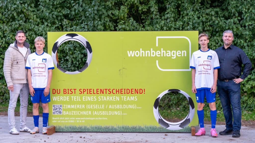 Vor einem grünen wohnbehagen-Schild mit Fußballmotiven stehen zwei Erwachsene in legerer Kleidung neben zwei jugendlichen Fußballspielern. Die Gruppe lächelt bei der Trikotübergabe Westfalia Gemen im Freien und vermittelt ein Gefühl von Gemeinschaft und Wohlbefinden.