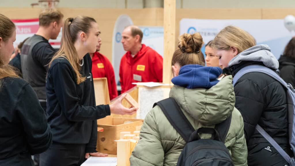 An einem gemütlichen Messestand aus Holz erläutert eine Frau drei Schülern die nachhaltigen Eigenschaften von Holzbausteinen. Die freundliche Atmosphäre und das moderne Design der Ausstellung laden ein, mehr über wohngesundes Bauen zu erfahren.
