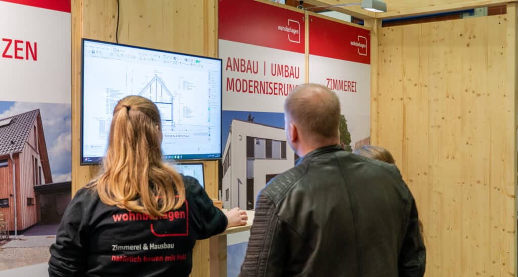 Drei Menschen stehen an einem Stand mit Holzverkleidung und informieren sich vor einem großen Bildschirm über nachhaltige Architekturpläne. Umgeben von Bannern zu Renovierung, Modernisierung und Zimmerei entsteht hier Wohlfühlatmosphäre rund ums Eigenheim.