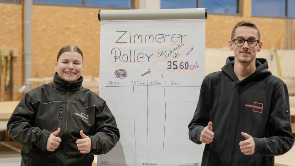 Zwei junge Menschen stehen in einer gemütlichen Werkstatt mit Holzmaterialien, lächeln und zeigen Daumen hoch vor einem Flipchart mit der Aufschrift "Zimmerer Rally" und bunten Zeichnungen - ein Moment voller Freude beim nachhaltigen Hausbau.