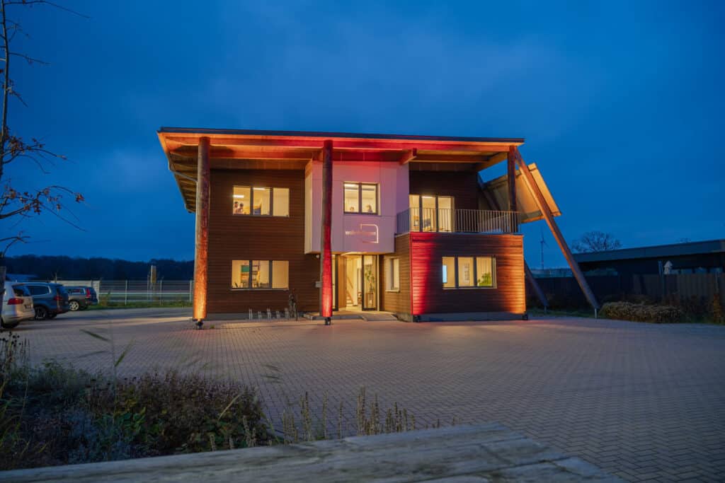 Ein modernes, zweistöckiges Holzhaus mit großen Fenstern strahlt dank warmer Außenbeleuchtung eine behagliche Atmosphäre bei einsetzender Dämmerung aus. Die natürliche Holzfassade und das geneigte Dach unterstreichen den wohnlichen Charakter. Vor dem Haus befindet sich ein gepflasterter Parkplatz mit wenigen Autos.