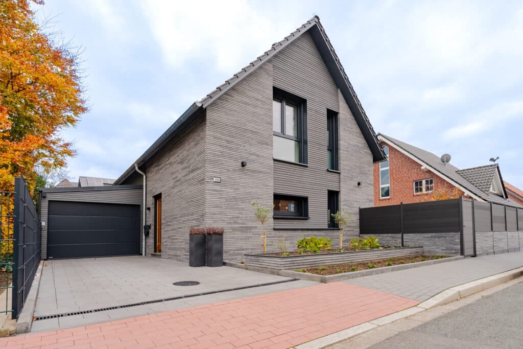 Modernes Haus in zeitgemäßem Design mit grauer Ziegelfassade, geneigtem Dach und großen Fenstern. Der liebevoll gestaltete Vorgarten und die natürliche Umgebung mit herbstlichen Bäumen schaffen ein behagliches Zuhause zum Wohlfühlen.