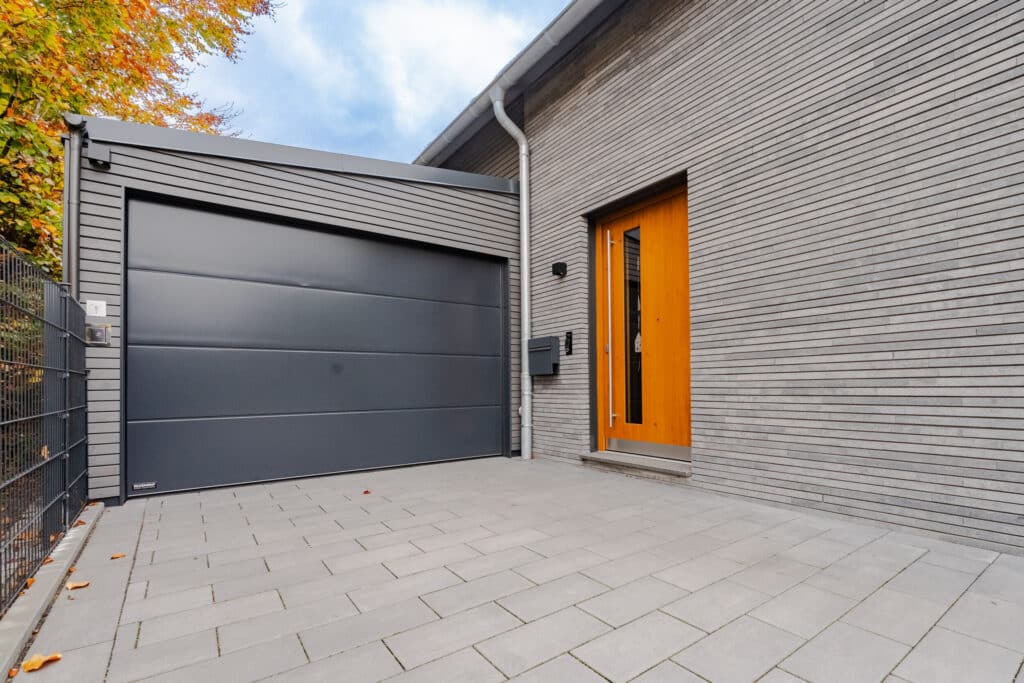 Ein modernes Haus mit grauen Ziegelwänden und großem dunklen Garagentor strahlt zeitlose Eleganz aus. Die Holzeingangstür sowie der mit hellen Fliesen gepflasterte Weg sorgen zusammen mit herbstlicher Bepflanzung für Wohnbehagen.