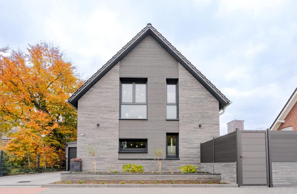 Ein modernes, graues Holzhaus mit steilem Dach und großen Fenstern steht einladend vor einem minimalistischen Garten. Natürliche Materialien, großzügige Glasflächen und ein Baum mit herbstlichen Blättern schaffen Behaglichkeit.
