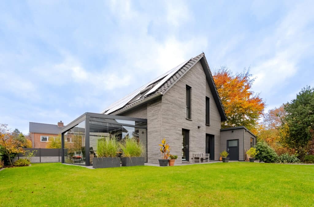 Modernes, zweistöckiges Haus mit grauer Klinkerfassade, großen Fenstern und Solarpanelen auf dem Dach. Die verglaste Terrasse lädt zum Wohlfühlen ein. Umgeben von grünem Rasen, Bäumen und Pflanzen strahlt das Zuhause Behaglichkeit aus.