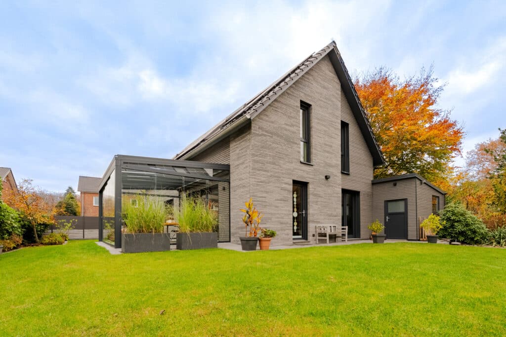 Modernes zweistöckiges Holzfertighaus mit lichtgrauer Fassade, großzügigen Fenstern und elegantem Pultdach. Umgeben von gepflegtem Rasen und herbstlichen Bäumen öffnet sich die Terrasse, liebevoll gestaltet mit Pflanzen und gemütlichen Sitzmöbeln - einladend zum Wohlfühlen.