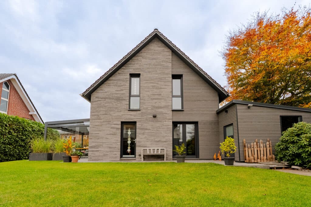 Ein modernes, zweistöckiges Holzhaus mit grauer Fassade, großen Fenstern und geneigtem Dach vermittelt zeitgemäßen Wohnkomfort. Es ist eingebettet in einen grünen Garten mit pikierten Pflanzen und leuchtend orangefarbenem Herbstlaub - eine Bank lädt zum Verweilen ein.
