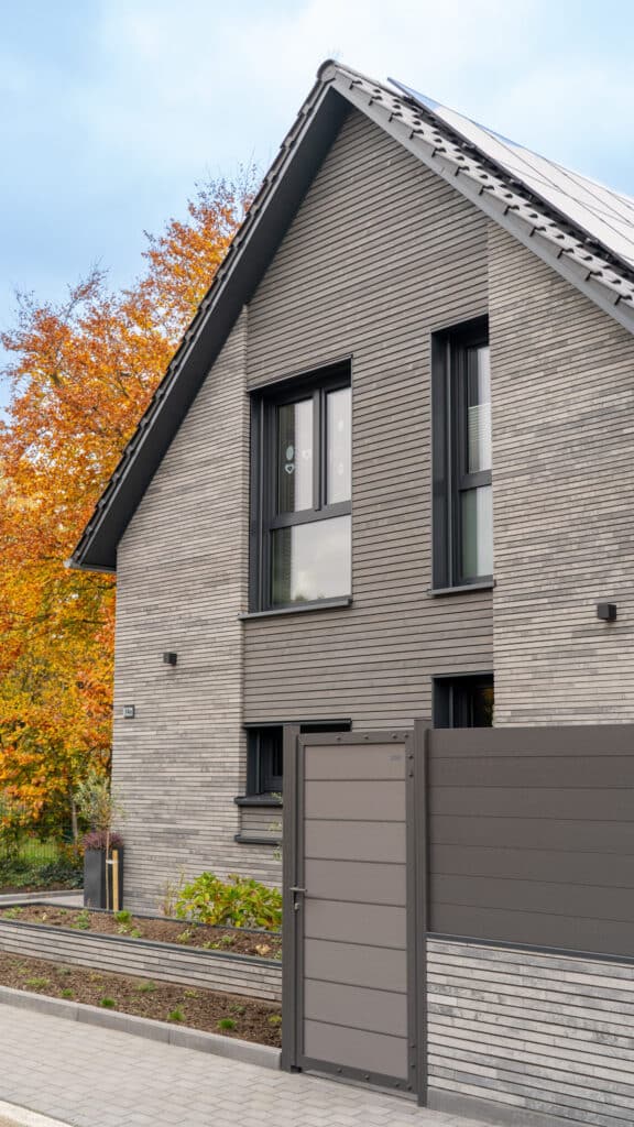 Modernes Holzfertighaus in elegantem Grau mit großen Fenstern, geneigtem Dach und passendem Zaun. Die natürliche Umgebung mit herbstlich gefärbten Bäumen sorgt für eine warme Atmosphäre, während der Himmel teils bewölkt ist.