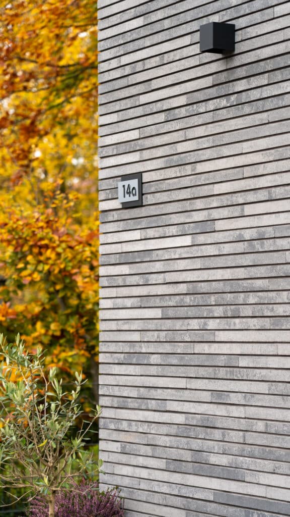 Eine moderne graue Ziegelwand mit der Hausnummer 14a und einer eleganten schwarzen Außenleuchte, eingerahmt von herbstlichen Bäumen und Pflanzen - ein harmonisches Zusammenspiel aus zeitgemäßem Design, natürlichen Materialien und wohnlicher Atmosphäre.