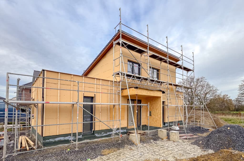 Ein modernes, zweistöckiges Holzhaus im Bau wird von Außengerüsten umgeben. Die Fassade ist noch nicht fertiggestellt, natürliche Materialien liegen bereit. Vor wolkenverhangenem Himmel und grünen Bäumen entsteht hier ein behagliches Zuhause.